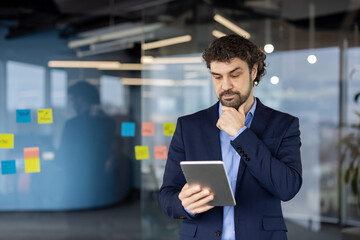 Businessman in blue suit deeply analyzes information on tablet. Thoughtful expression highlights focus on data-driven decision making in modern office setting with colorful sticky notes.