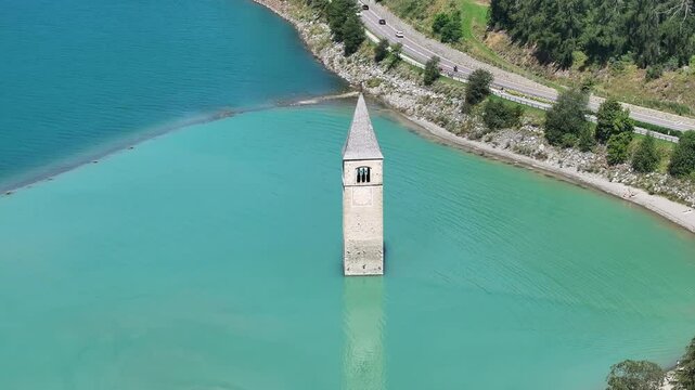 An aerial view of the submerged bell tower of Curon Venosta (Graun im Vinschgau) rising from Lake Reschen in South Tyrol, Italy. A unique glimpse into the region's history and natural beauty.