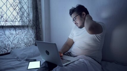 Indian young man using laptop and overworking at night on bed, having pain in neck. - Powered by Adobe