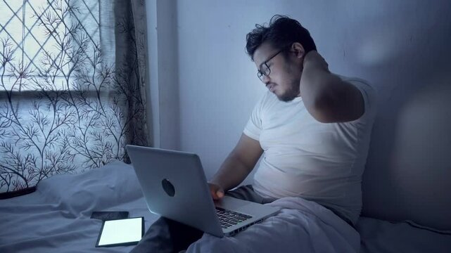 Indian young man using laptop and overworking at night on bed, having pain in neck.