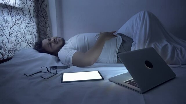A man lying on the bed suffering from severe stomach pain, diarrhoea, he overworking with computer technology during night time.