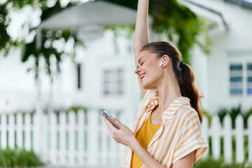 Young woman enjoying music outdoors, smiling joyfully with arm raised, wearing wireless earphones,...
