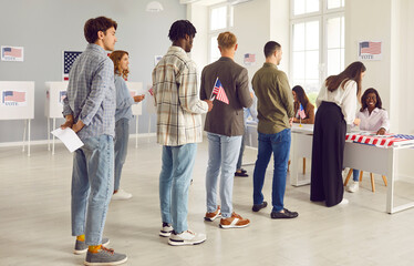 Naklejka premium Large group of diverse smiling people registering at polling station holding American flags in hands on election day. American voters standing in queue at vote center among citizens of USA.