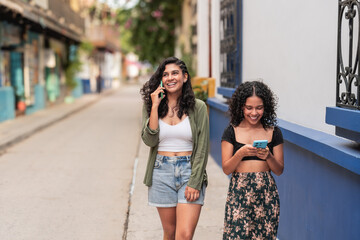 Two women walking outdoors while using smartphones on a sunny street.