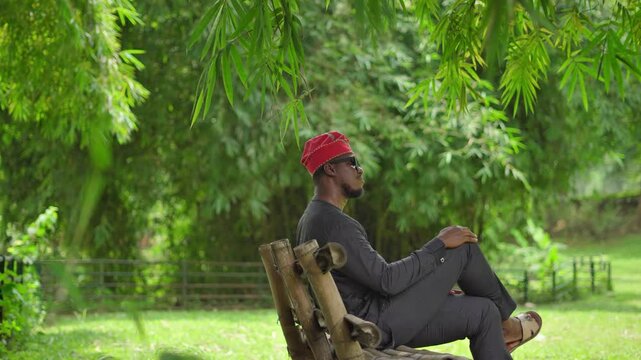 Young African Yoruba male sitting thoughtfully in a garden, crosses his legs.
