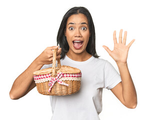 Latina holding a wicker basket receiving a pleasant surprise, excited and raising hands.