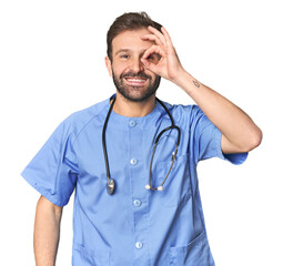 Hispanic male nurse in studio excited keeping ok gesture on eye.