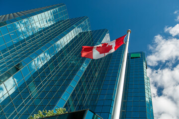 Canadian Flag waves in front of a building in Ottawa, clouds and blue sky reflections on the building windows. © PAOLO