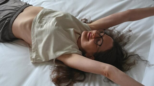 Young woman lying on a bed, stretching her arms and smiling, radiates relaxation and comfort in a cozy bedroom setting