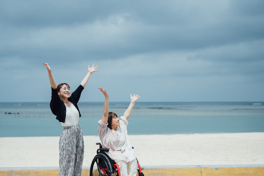 車椅子に乗った日本人女性が友人と手を広げ沖縄の海の開放感を感じている