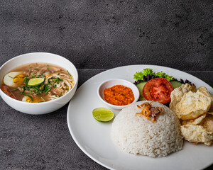 A plate of steamed rice with sambal, kerupuk, vegetables, and a bowl of chicken soup with egg, lime, and herbs on the side.
