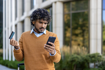 Man with curly hair wearing brown sweater holding credit card and smartphone. Standing outdoors appearing confused, possibly facing online banking or payment problem during business trip.