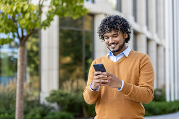 Young professional with curly hair using smartphone outdoors. Scene conveys connectivity, technology, and communication. Individual dressed in casual autumn attire, standing confidently near office