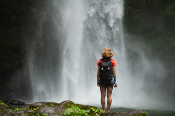 Obraz premium A young blonde girl, a traveler, admires a majestic waterfall with a backpack.
