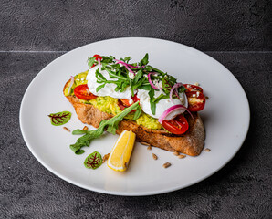 Avocado toast garnished with poached eggs, cherry tomatoes, arugula, red onion slices, and a lemon wedge, served on a crispy slice of bread.