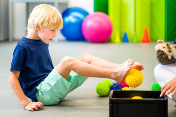 Child during physical therapy session. Boy exercising barefoot with textured surface ball in physio...