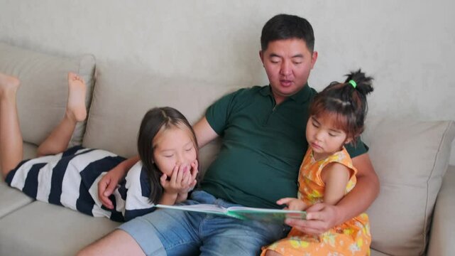 Two cute girls reading a book with fairy tales sitting on the couch with their father, time together in asian family.