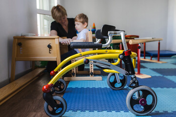 Cute boy with neurodevelopmental disorders is participating in a rehabilitation session under guidance of therapist in therapy space with rehab walker