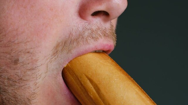 Close up - Unrecognized hungry man takes big bite of hot dog on gray background. Concept of junk food, fast food, which will subsequently cause problems of the digestive system and digestion.