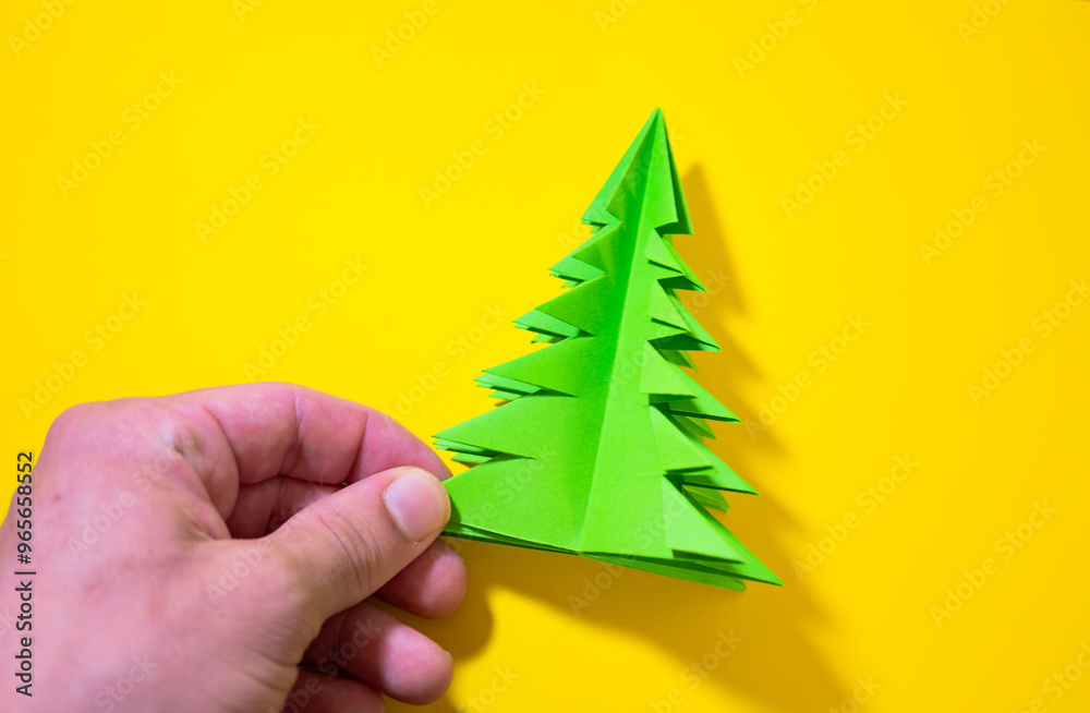 Wall mural male hand holding a green tree made in origami christmas concept on a yellow background