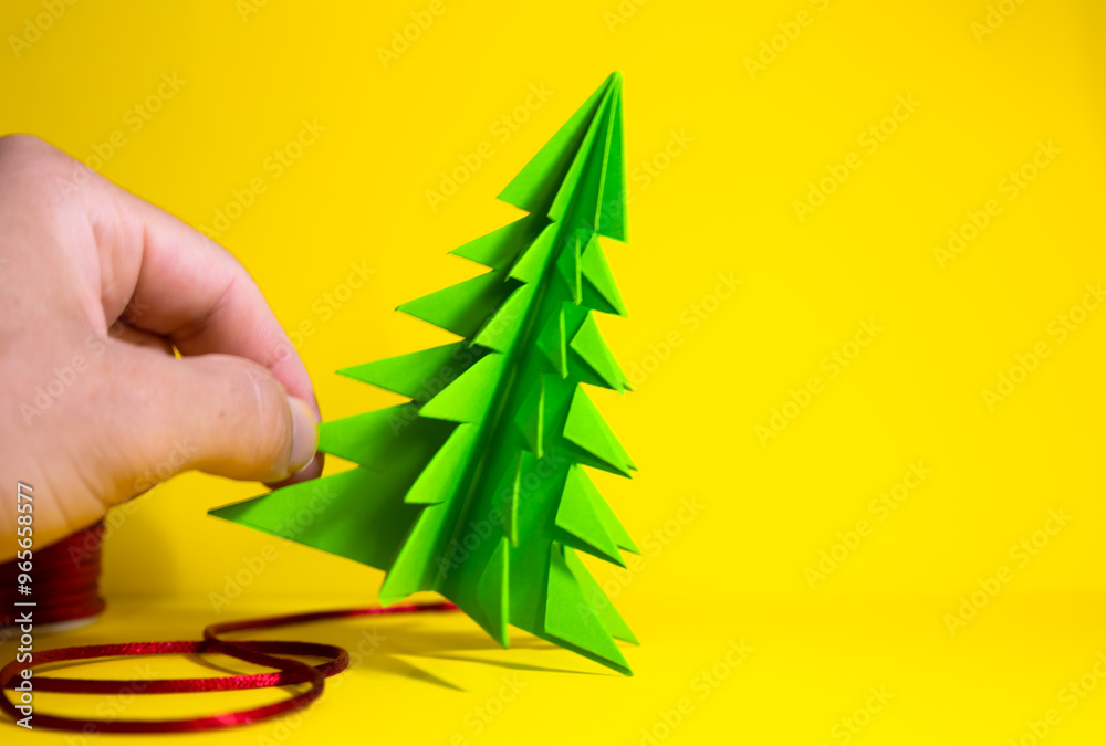 Wall mural male hand placing a christmas paper tree on a yellow background, christmas concept and decoration