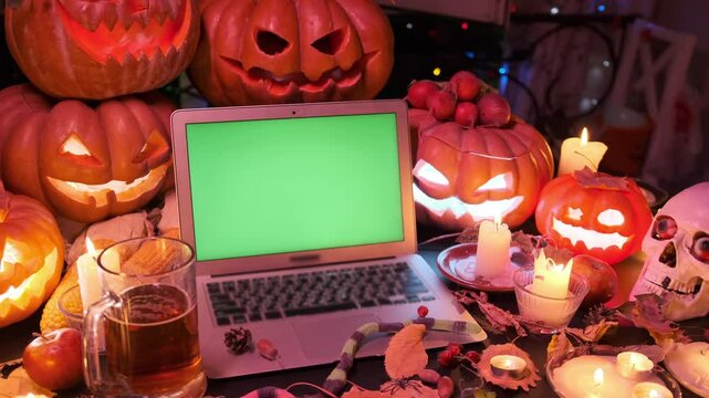 Happy Halloween night party offers. Laptop with green chromakey on screen and many glow jack o'lanterns, candles, pumpkins and mug of beer, holiday background, close up.