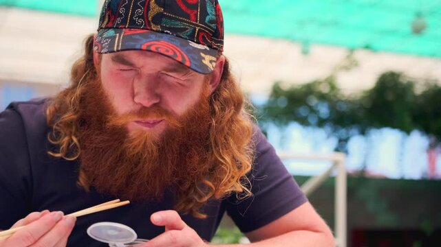 Curly man with red beard in cap eats delivery food japanese sushi rolls wincing from the spiciness of wasabi sitting at table on street. Close up.