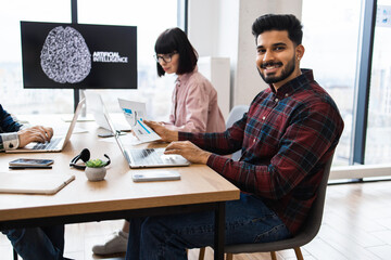 Team working together on artificial intelligence project in modern office setting. Smiling team member looking at camera while others focus on laptops and documents. High-tech work environment.