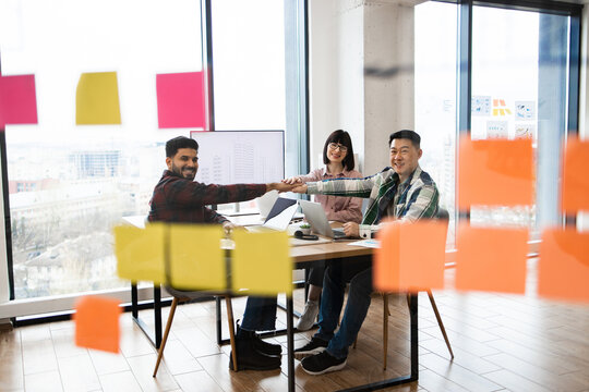 Multicultural team of architects collaborating on project in bright office. Three people sitting around table with laptops, sticking notes visible on glass wall, sharing ideas and shaking hands.
