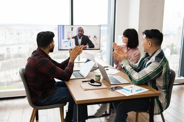 Business team engaging in conference call with colleague, expressing success and appreciation through applause, sitting around meeting table with laptops and documents, having productive work session.