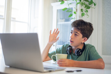 A young boy greets the people in a video conference he is holding on a laptop. He is holding a cup...