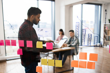 Business team having meeting with charts and post-it notes. Man using laptop with colleagues in background discussing business strategy and data analysis.