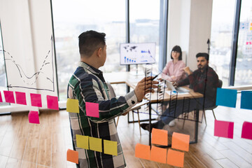 Business meeting with team brainstorming on charts and sticky notes. Colleagues discussing strategy in modern office space. Man presenting ideas and graphs to team members on glass wall.