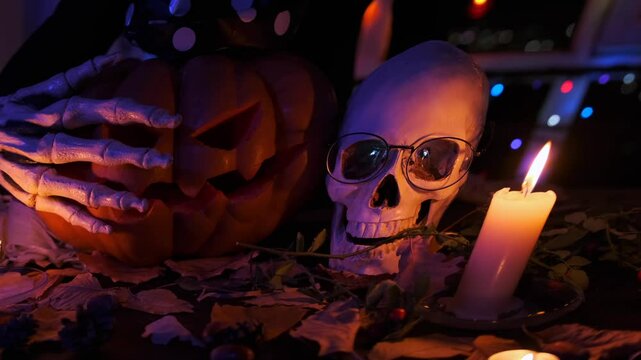 Close up, skeleton terrible paws touching pumpkins Jack Lantern, brightly flashing inside near human skull, on Halloween party, spooky invitation background.