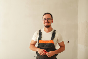 A construction worker dressed in overalls smiles confidently while standing against a white wall. The clean background emphasizes the worker's friendly demeanor and professional appearance