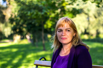 A portrait of a woman in her 40s, with blonde hair, wearing a purple blouse, set against a nature-filled park background. The image focuses on her face, capturing a moment of calm and contentment