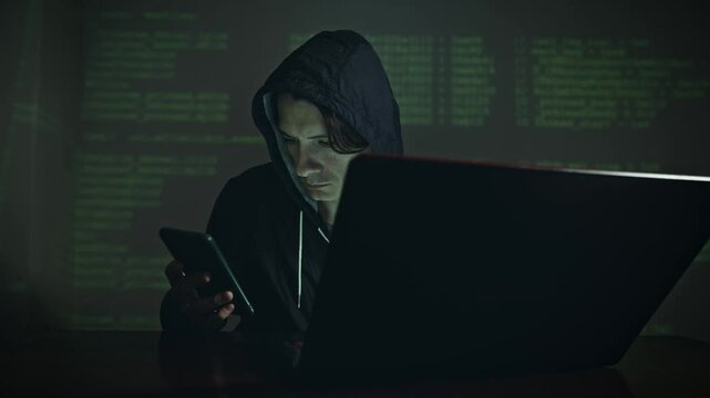 Male hacker in the hood holding the phone in his hands trying to hack the mobile device cloud and steal data in neon light. The concept of cyber security