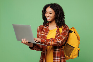 Young happy smiling teen girl IT student wear brown yellow casual clothes backpack bag use work on laptop pc computer isolated on plain green color background. High school university college concept.