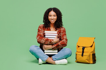 Full body young smiling teen girl student wear brown yellow casual clothes backpack bag sits hold pile of books look camera isolated on plain green background. High school university college concept.