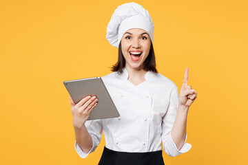 Young chef cook baker woman wear white shirt apron uniform toque chefs hat hold use digital tablet pc computer look for recipe point finger up isolated on plain yellow background Cooking food concept