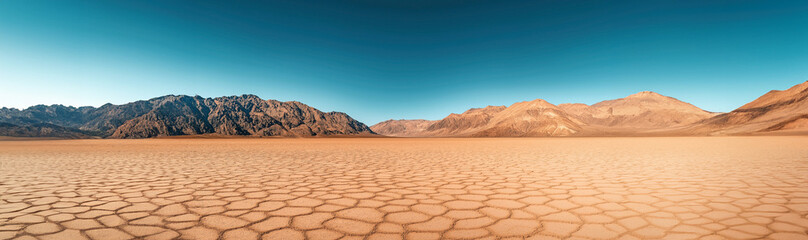 Wide-angle view of a dry, cracked desert landscape with mountainous terrain in the background under a clear blue sky, emphasizing arid and barren conditions.