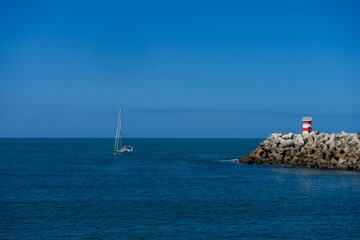 A boat approaches the entrance of Nazaré Marina, Portugal, with the lighthouse towers on both piers clearly visible, against a backdrop of calm waters and clear skies.
