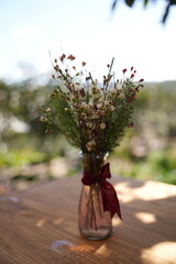 A bouquet of white and pink flowers tied with a red ribbon in a clear glass vase on a wooden table, outdoors with blurred greenery background.