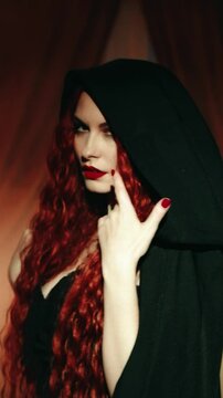Happy beauty halloween woman vampire gothic sexy queen looking back. Dark night room. Fantasy girl evil goth smiling face red lips long hair black dress hood on head old style vertical video crop art