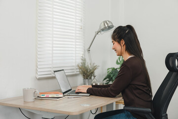 A woman is sitting at wooden desk, working on laptop with smile. workspace is bright and organized, featuring lamp, notebook, and cup, creating productive atmosphere.