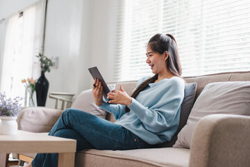 Obraz premium A young woman is sitting comfortably on sofa, engaging with tablet while smiling, creating warm and inviting atmosphere.