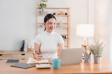 A young woman is smiling while working on her laptop at cozy home office. She is taking notes and appears focused and happy in her bright, well organized workspace.