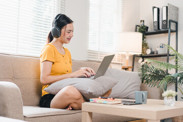 A woman wearing headphones is sitting comfortably on sofa, working on her laptop with smile. cozy atmosphere is enhanced by natural light and stylish decor.