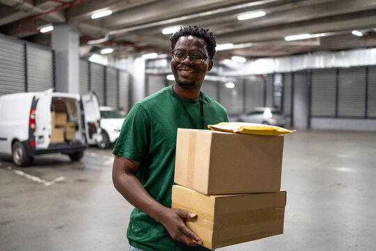 Experienced black deliveryman working in packages sorting center, carrying boxes in the van.