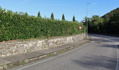 Yield sign at the end of the street, on concrete sidewalk. By side, a fence made of low stone wall...